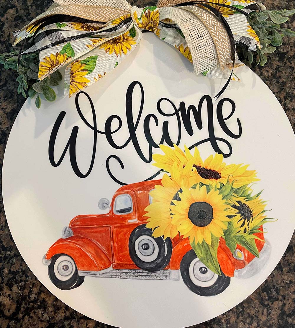 Large round home decor personalized with a pickup hauling large sunflowers, "Welcome", and a ribbon at the top