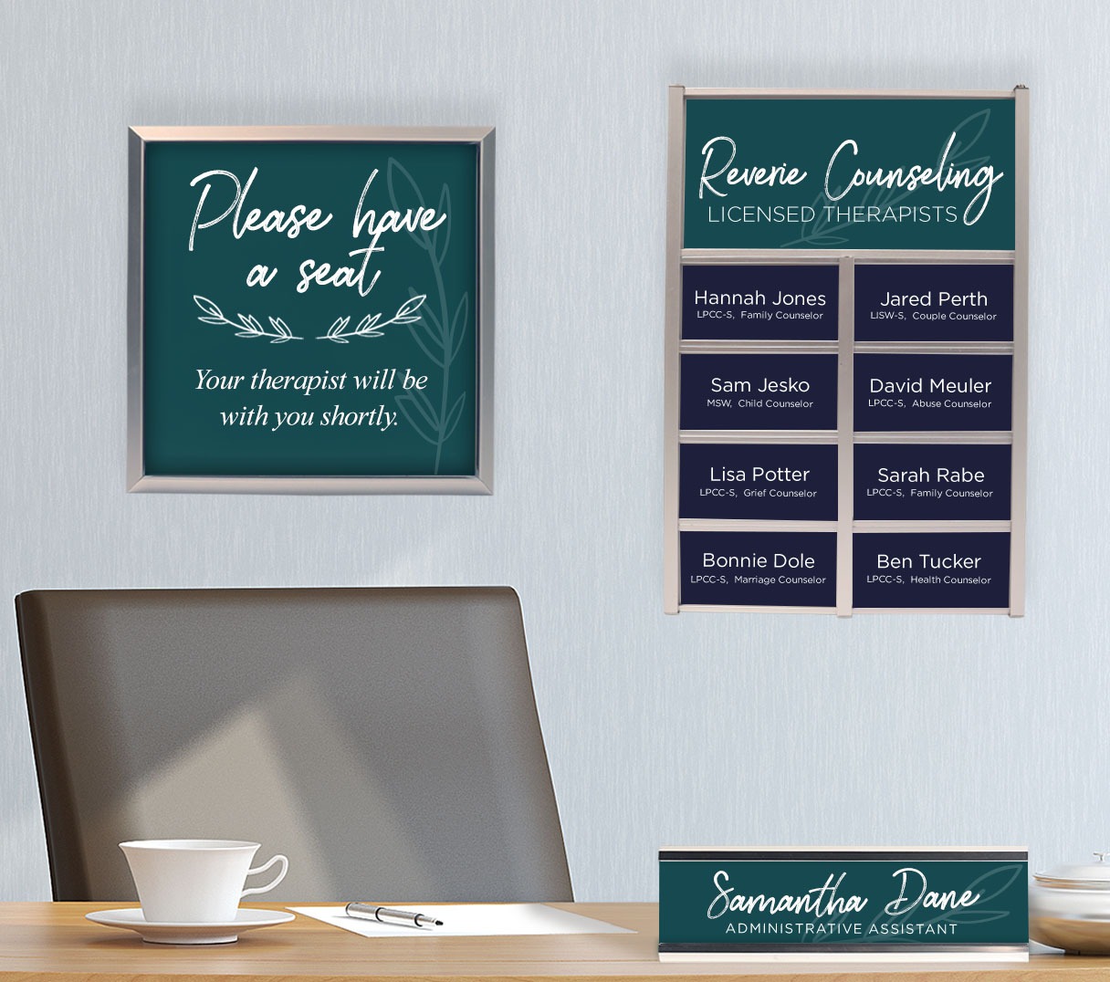 A desk with a tea cup, paper and pen, desk name plate, and on the wall behind a framed image to please have a seat next to mounted wall plate holder list of names