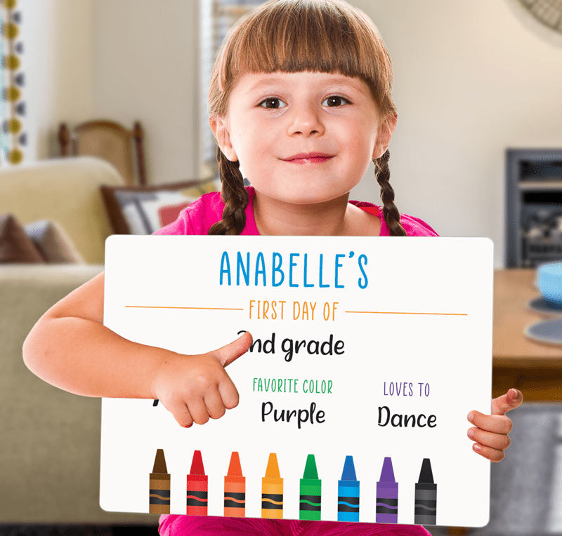 Young girl holding a sign for her first day of 2nd grade with her name on it images of crayons along the bottom